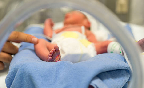 Photo of a premature baby in incubator. Focus is on his feet and toes. The doctor is touching him to check his reflexes. There are cables and tubes in the out-of-focus area.