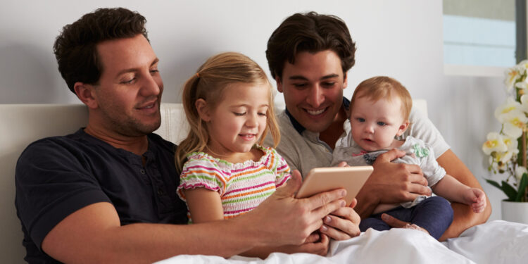 Male gay parents using tablet computer in bed with two kids