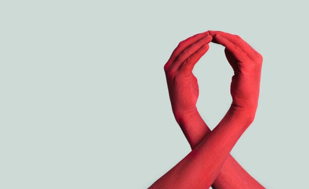 closeup of the arms of two men painted red forming a red awareness ribbon for the fight against AIDS, against an off-white background with some blank space on the left