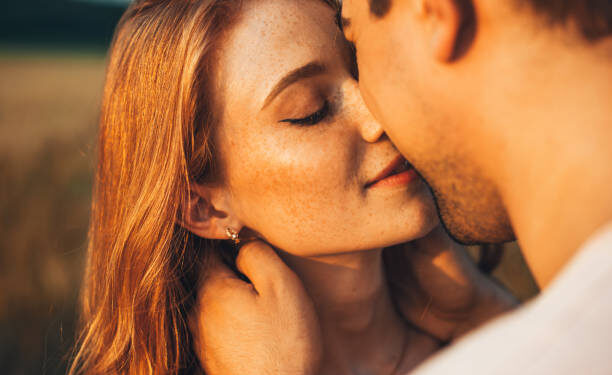 Close-up portrait of a freckled girl kissing her boyfriend while they are on an outdoor date. Wheat field. People lifestyle concept.