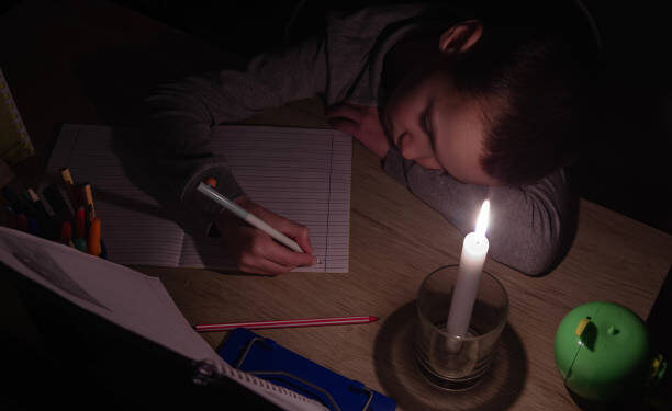 Tired schoolboy with candle in complete darkness doing homework. Power outage, blackout, concept image.