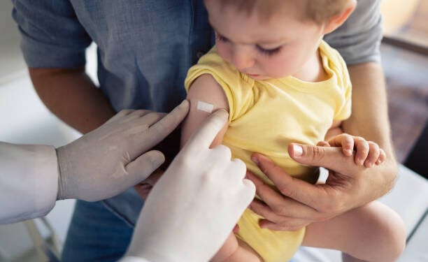 Unrecognizable doctor putting a patch on the little boy's shoulder after successful vaccination. Little boy sitting in father´s lap and looking at the patch.