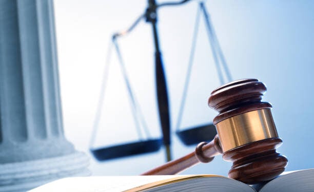 A gavel rests on top of an open law book with a justice scale and a Greek column in the background.