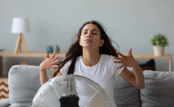 Funny overheated woman enjoying fresh air, cooling by electric fan, exhausted young female waving hands, sitting on couch at home in front of ventilator, suffering from hot summer weather