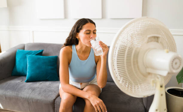 Happy woman trying to cool down drinking an ice cold drink and turning on the electric fan at home