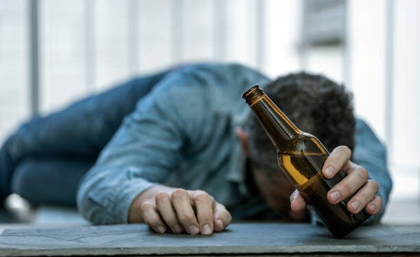 DRUNK MAN LYING ON THE FLOOR ASLEEP WITH A BOTTLE OF BEER IN HIS HAND. ALCOHOL CONSUMPTION ADDICTION. ALCOHOLISM CONCEPT. FOCUS SELECTED.