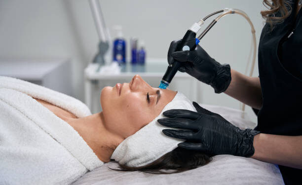 Cosmetologist in protective gloves sitting near beautiful lady and doing Hydradermabrasian procedure in salon