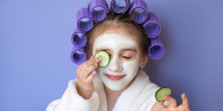 A little lady with a cleansing clay mask on her face and cucumber slices. Curls wrapped in curlers.  Daily care to maintain beauty and youth